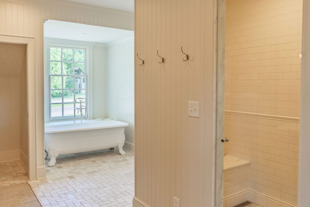 Hallway with hooks connecting a bedroom closet area to a remodeled primary bathroom with a freestanding tub